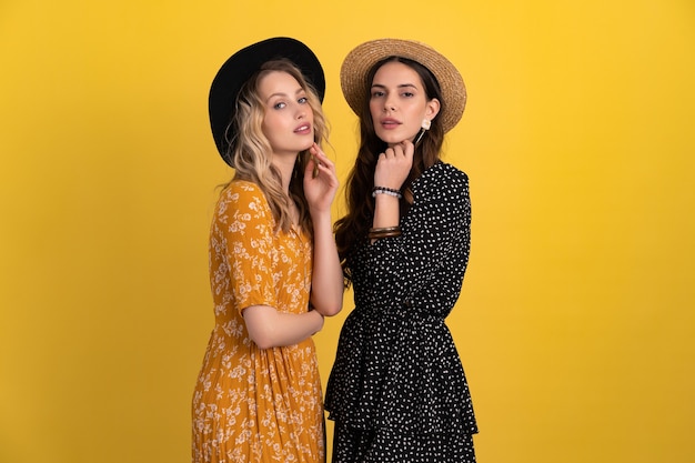 Две молодые красивые женщины-друзья вместе изолированы на желтом фоне в черно-желтом платье и шляпе