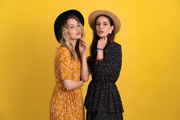 Two young beautiful women friends together isolated on yellow background in black and yellow dress and hat