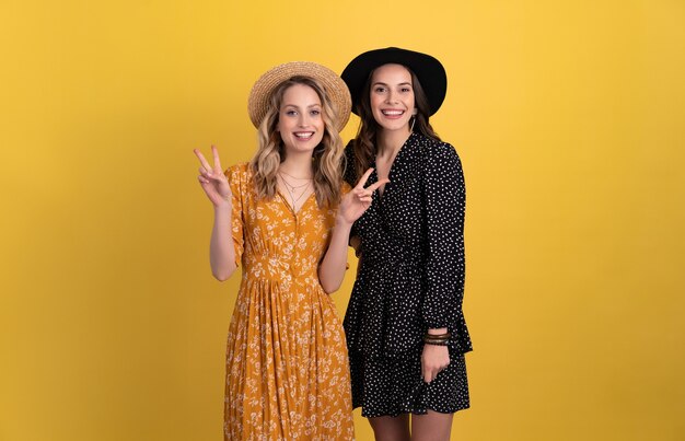 two young beautiful women friends together isolated on yellow in black and yellow dress and hat stylish boho having fun