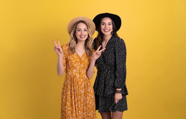 two young beautiful women friends together isolated on yellow in black and yellow dress and hat stylish boho having fun