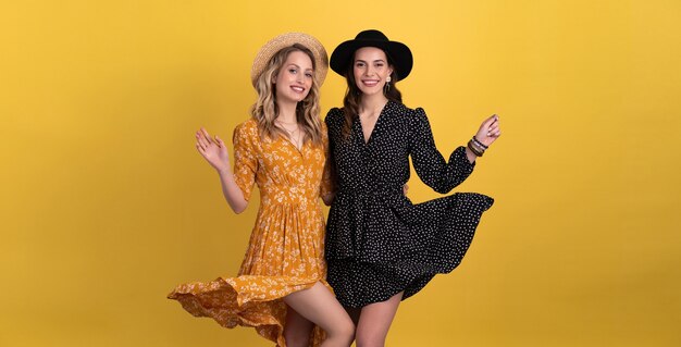 two young beautiful women friends together isolated on yellow in black and yellow dress and hat stylish boho having fun