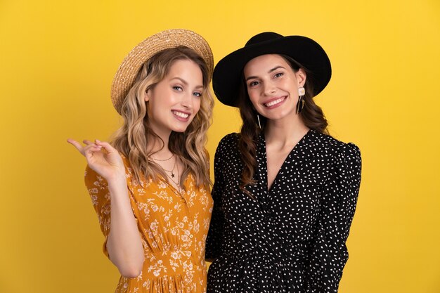 two young beautiful women friends together isolated on yellow in black and yellow dress and hat stylish boho having fun