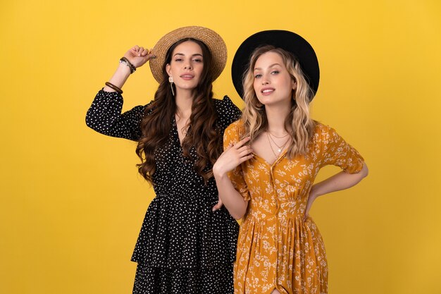 two young beautiful women friends together isolated on yellow in black and yellow dress and hat stylish boho having fun