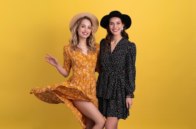 two young beautiful women friends together isolated on yellow in black and yellow dress and hat stylish boho trend
