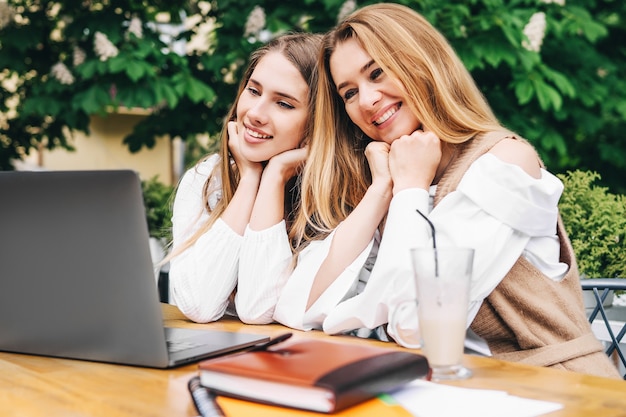 Two young blonde women are sitting at a table and looking at the computer with a smile