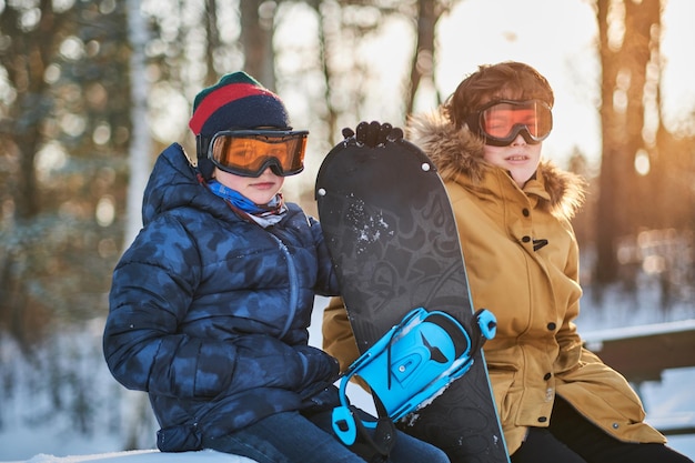 Two young boys are enjoying winter walk at forest with snowboard.