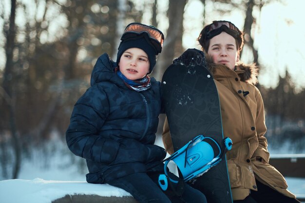 2人の少年がスノーボードを持って森で冬の散歩を楽しんでいます。