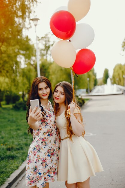 two young and bright girls spend their time in the summer park with balloons and phone