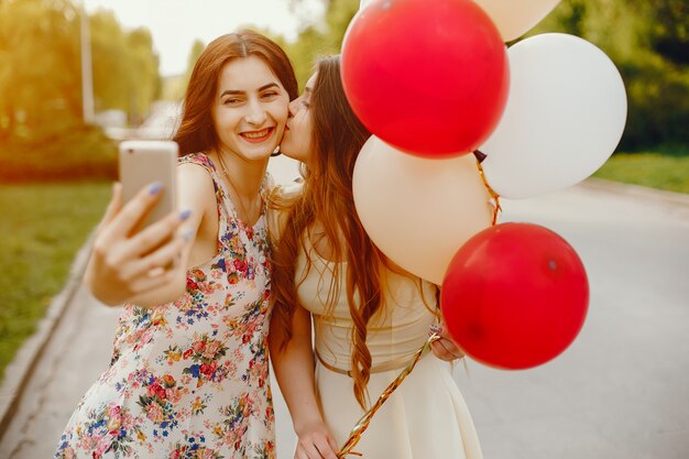 two young and bright girls spend their time in the summer park with balloons and phone