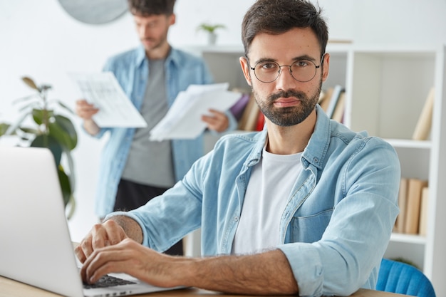 Two young businessmen spend productive morning in office, develop company`s strategy, work with laptop computer and business papers