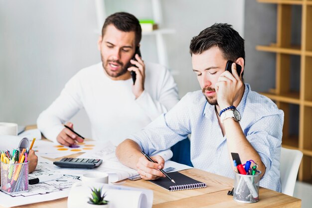 Two young businessmen talking on smartphone at workplace