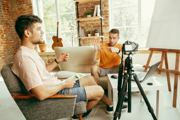 Two young caucasian male bloggers in casual clothes with professional equipment or camera recording video interview at home. Blogging, videoblog, vlogging. Talking while streaming live indoors.