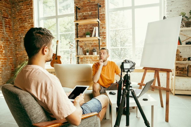 Two young caucasian male bloggers in casual clothes with professional equipment or camera recording video interview at home. Blogging, videoblog, vlogging. Talking while streaming live indoors.