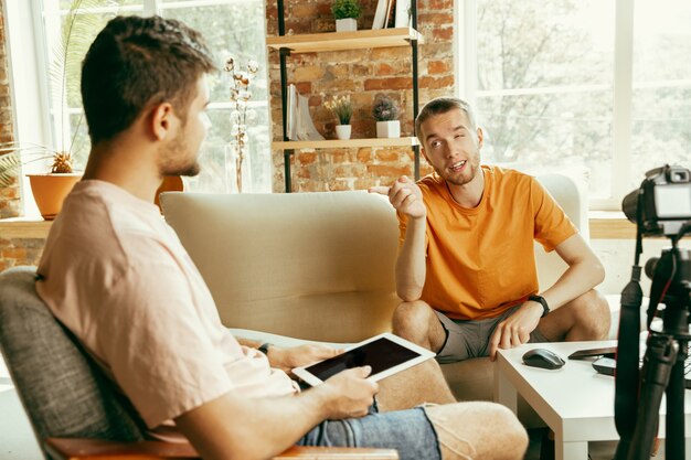 Two young caucasian male bloggers in casual clothes with professional equipment or camera recording video interview at home. Blogging, videoblog, vlogging. Talking while streaming live indoors.
