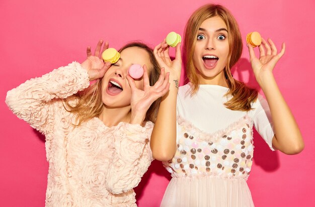 Two young charming  beautiful smiling hipster women in trendy summer clothes. Women making glasses, spectacles with colorful macaroons, holding macarons on eyes place. Posing on pink wall