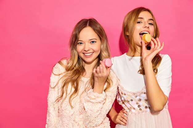 Two young charming  beautiful smiling hipster women in trendy summer clothes. Women  with colorful macaroons, holding macarons near face. Posing on pink wall