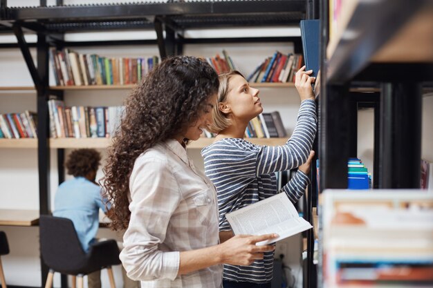 チームプロジェクトの文学を探している大学図書館の本棚の近くに立っているカジュアルな服装で2人の若い陽気な女子学生