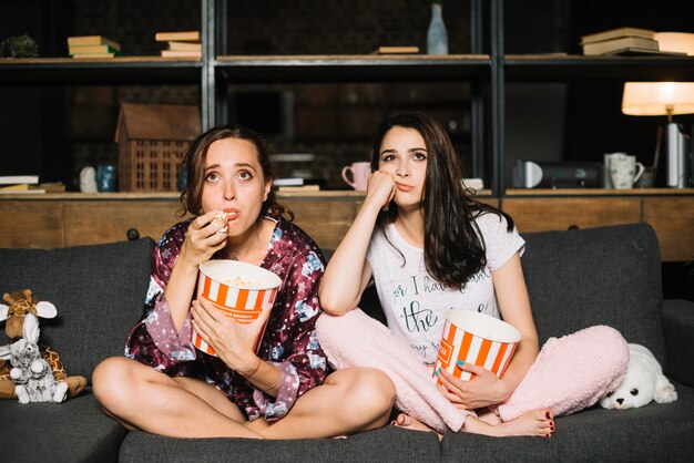 Две молодые женщины-друзья, сидящие на диване, смотрят телевизор