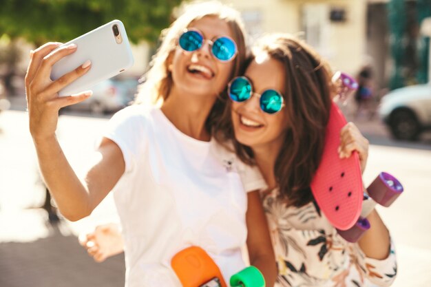 Two young female stylish hippie brunette and blond women models in summer hipster clothes taking selfie photos for social media on smartphone