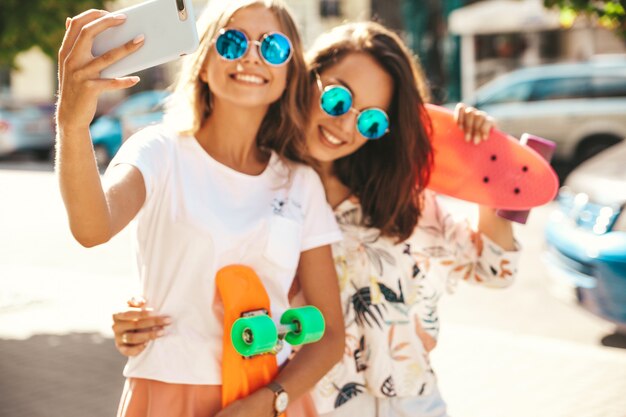 Two young female stylish hippie brunette and blond women models in summer hipster clothes taking selfie photos for social media on smartphone