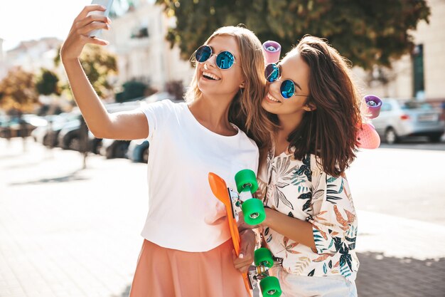 Two young female stylish hippie brunette and blond women. Models in summer sunny day in hipster clothes taking selfie photos for social media on phone . With colorful penn