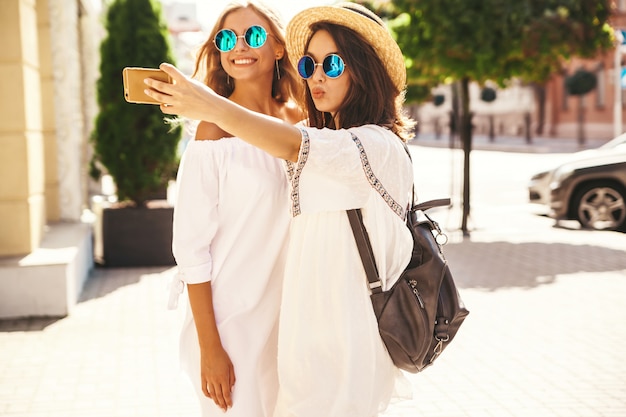 2人の若い女性のスタイリッシュなヒッピーブルネットとブロンドの女性は、電話でソーシャルメディアのselfie写真を撮る白い流行に敏感な服で夏の晴れた日にモデルします。