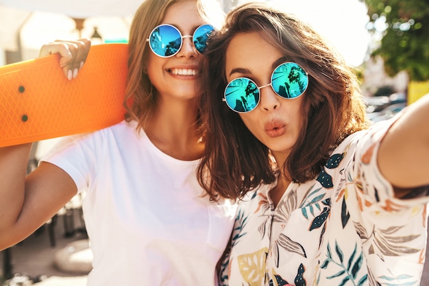 Due giovani donne alla moda hippy brunette e donne bionde in pantaloni a vita bassa estate vestiti prendendo selfie