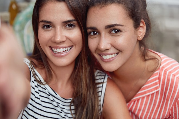 Two young females take selfie with unrecognizable device, have broad smiles, white perfect teeth, spend free time together, being in good mood. Pretty brunette woman makes photo as stands with friend