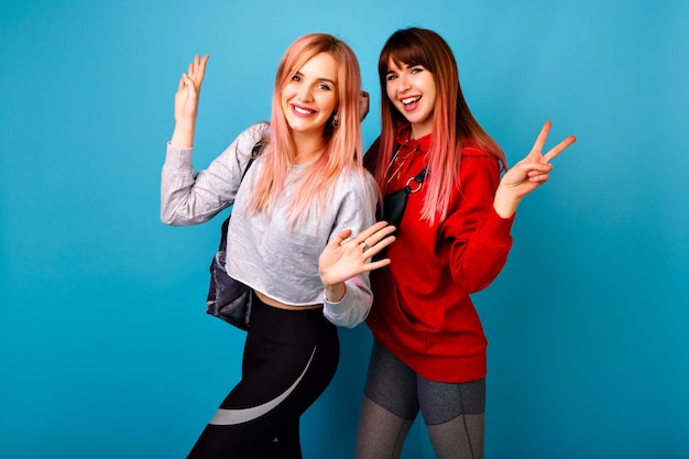 Две молодые забавные симпатичные хипстерские женщины в ярких спортивных костюмах, улыбаются, кричат и здороваются, синяя стена