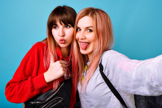 Two young funny pretty hipster women wearing sportive bright casual outfits, smiling screaming and say hi to you, blue  wall