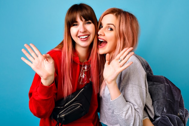 Two young funny pretty hipster women wearing sportive bright casual outfits, smiling screaming and say hi to you, blue  wall