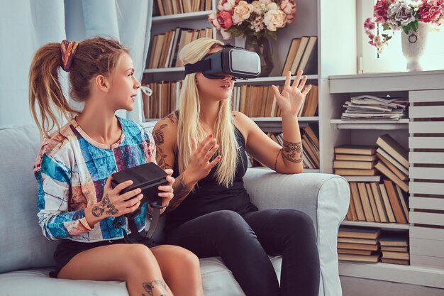 Two young girlfriends in casual clothes having fun with virtual reality glasses device at home.