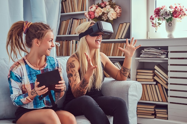 Two young girlfriends in casual clothes having fun with virtual reality glasses device at home.