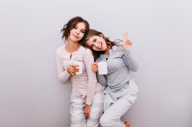 Two young girls in pajamas with cups on gray wall . Girl with long hair lean head on shoulder of girl with curly hair. They smiling .