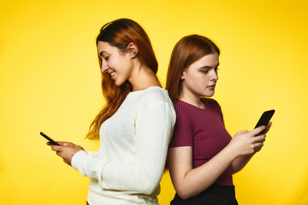 Две молодые девушки стоят спиной к спине и смотрят в свои телефоны стоя