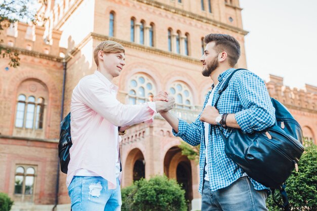 Два молодых симпатичных парня студентов с рюкзаками приветствуют друг друга на территории кампуса. в университете.