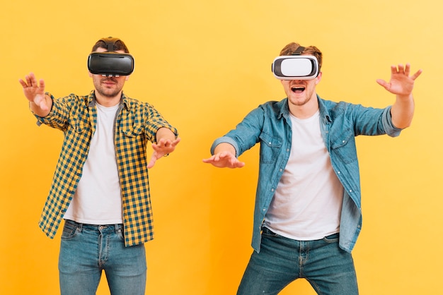 Two young man using virtual reality goggles touching in the air against yellow background