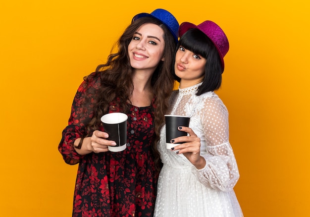 플라스틱 커피 컵을 들고 오렌지색 벽에 격리된 다른 입술을 헐떡이며 웃고 있는 파티 모자를 쓴 두 명의 젊은 파티 소녀