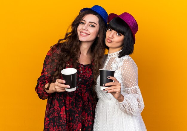 Two young party girls wearing party hat both holding plastic coffee cup one smiling another pursing lips isolated on orange wall