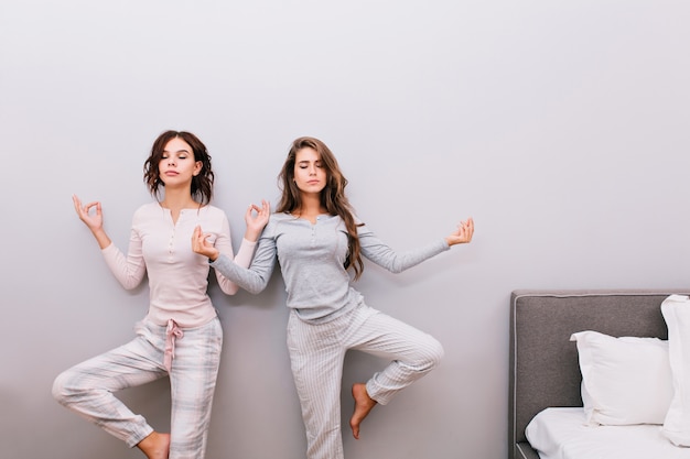 Two young pretty girls in night pajamas on grey wall . They doing meditation with closed eyes.