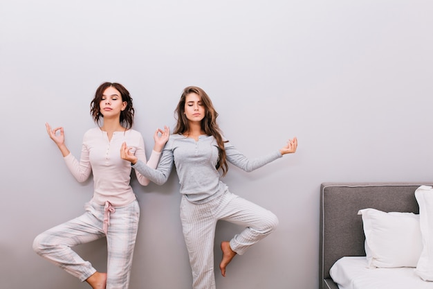 Две молодые красивые девушки в ночной пижаме на серой стене. Они медитируют с закрытыми глазами.