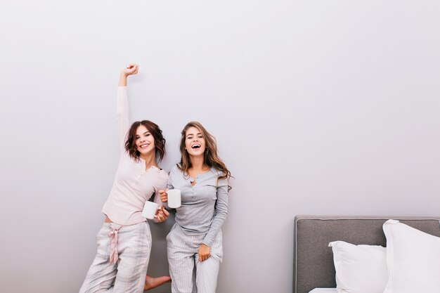 Two young pretty girls in pajamas with cups in sleeping room on grey wall . They having fun and smiling .