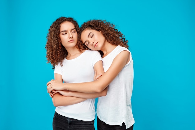 Two young pretty girls twins embracing, smiling over blue wall