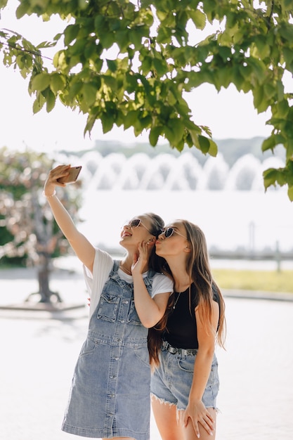 Two young pretty girls on a walk in the park taking pictures of themselves on the phone