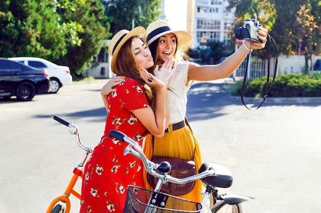 Due giovani ragazze abbastanza alla moda che posano vicino alle biciclette retro luminose dei pantaloni a vita bassa e che fanno autoritratto