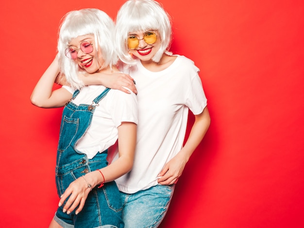 Two young sexy hipster girls in white wigs and red lips.Beautiful trendy women in summer clothes.Carefree models posing near red wall in studio summer  in sunglasses