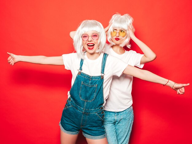 Two young sexy hipster girls in white wigs and red lips.Beautiful trendy women in summer clothes.Carefree models posing near red wall in studio summer  in sunglasses