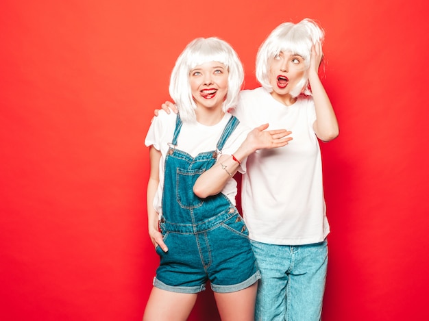 Две молодые сексуальные хипстерские девушки в белых париках и красных губах. Красивые модные женщины в летней одежде. Беззаботные модели позируют возле красной стены в студии летом в солнцезащитных очках