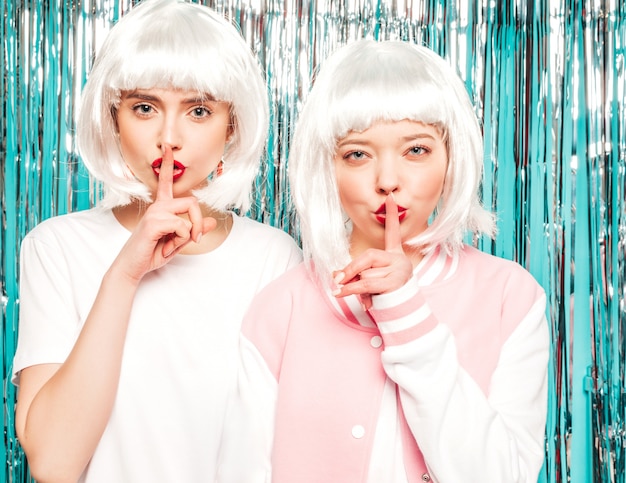 Two young sexy hipster girls in white wigs and red lips.beautiful trendy women in summer clothes.models posing on blue silver shiny tinsel background in studio.showes finger hush silence sign, gesture