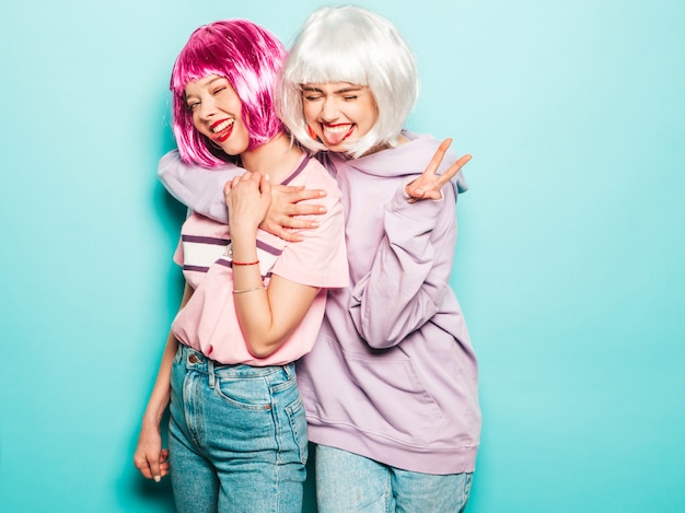 Two young sexy hipster girls in  wigs and red lips.Beautiful trendy women in summer clothes.Carefree models posing near blue wall in studio going crazy.Shows tongue